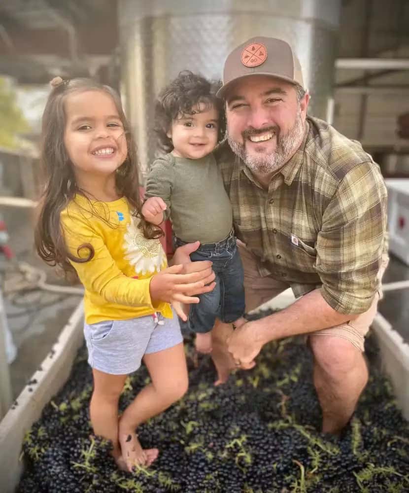 Smiling father with two young children standing barefoot in a vat of freshly harvested grapes, enjoying a family winemaking experience at a vineyard.