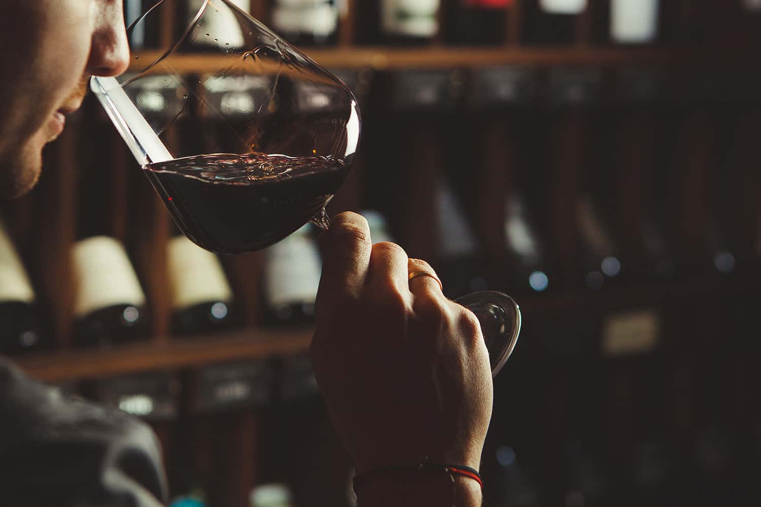 Man evaluating red wine in a glass at a dimly lit wine cellar, with shelves of wine bottles in the background.