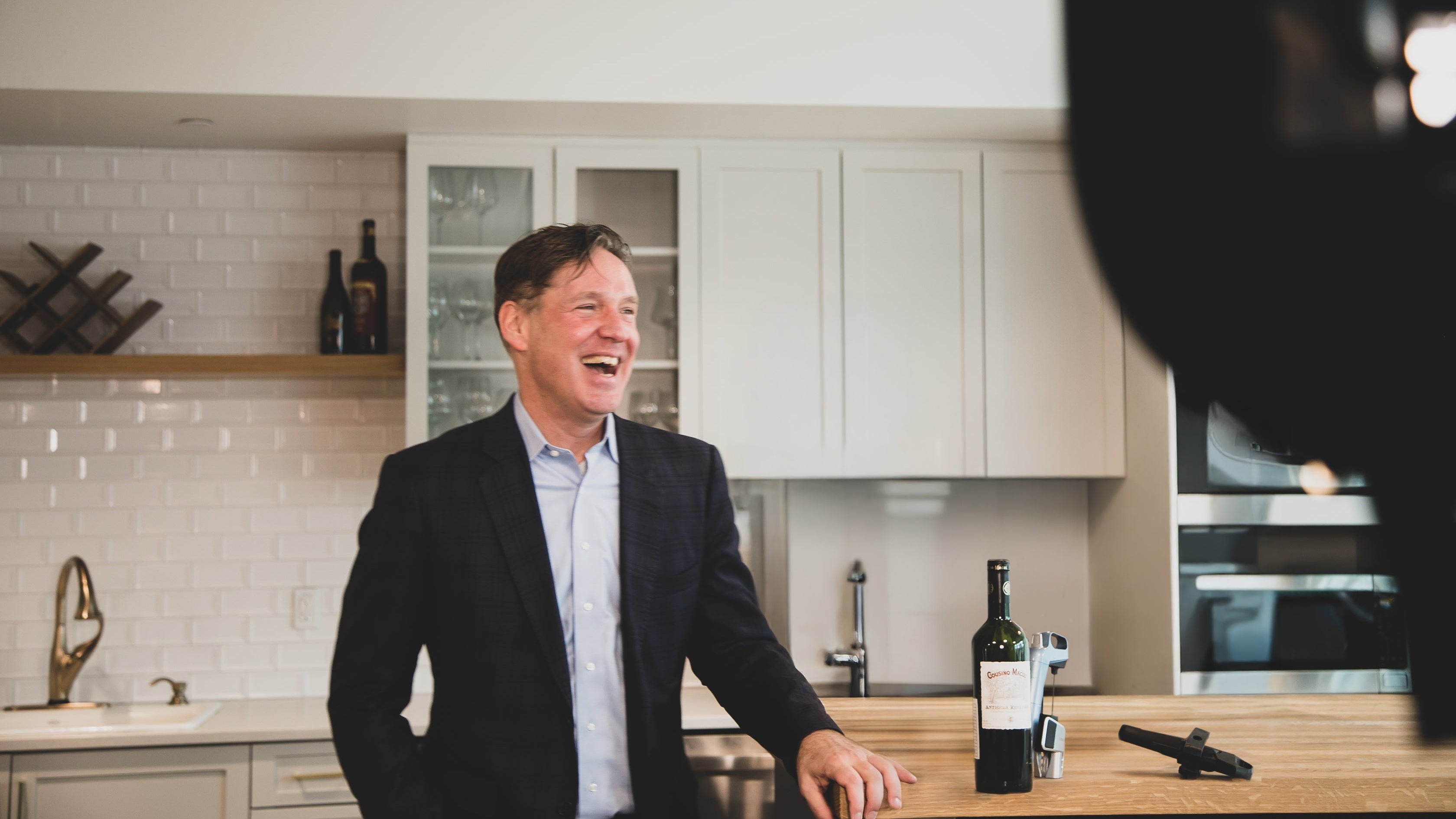 A man in a suit laughs while standing at a kitchen counter with a bottle of wine and a wine opener. Modern kitchen backdrop.