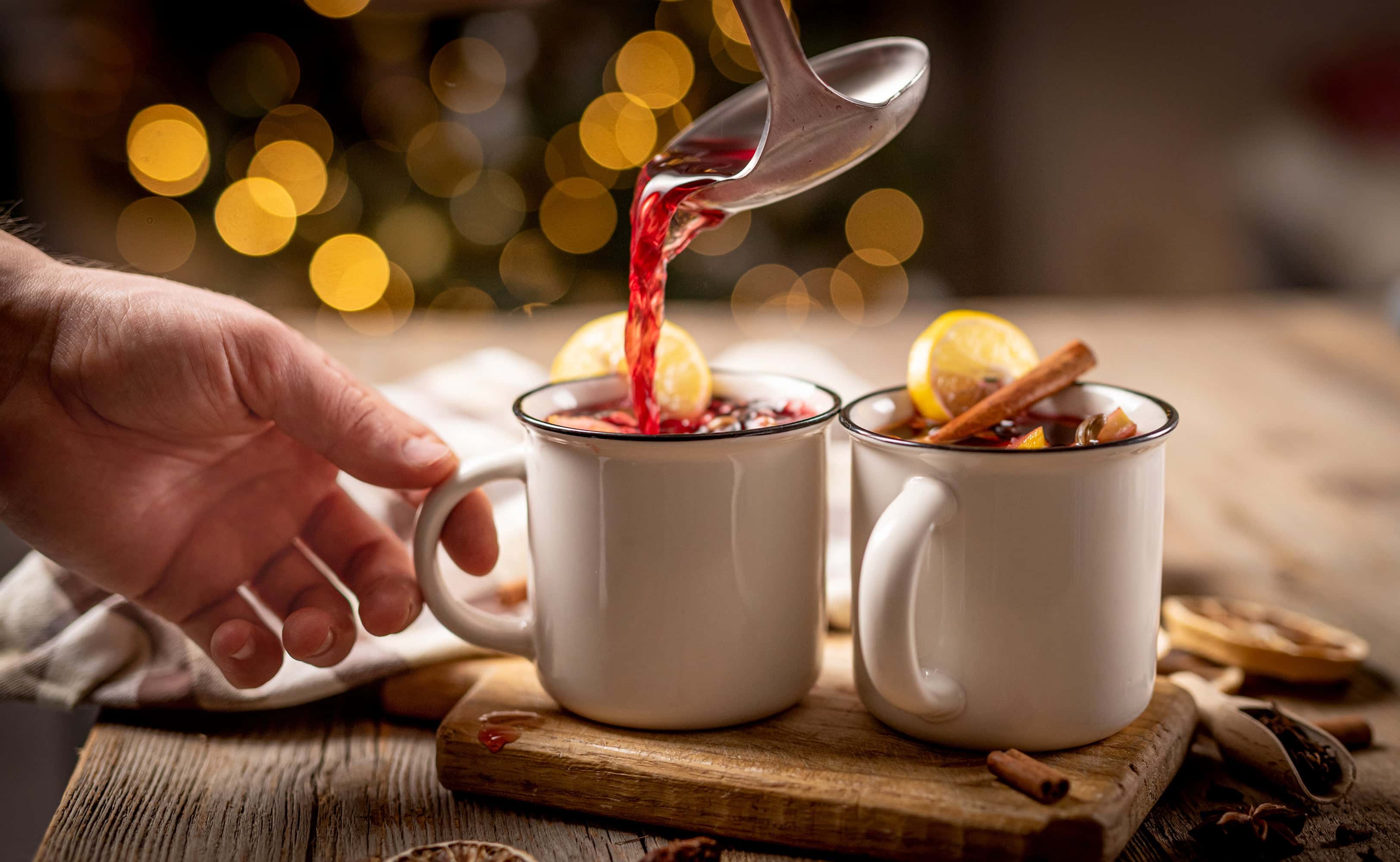 Hand pouring mulled wine into festive mugs with lemon slices and cinnamon sticks, set on a rustic wooden table with holiday bokeh lights in the background.