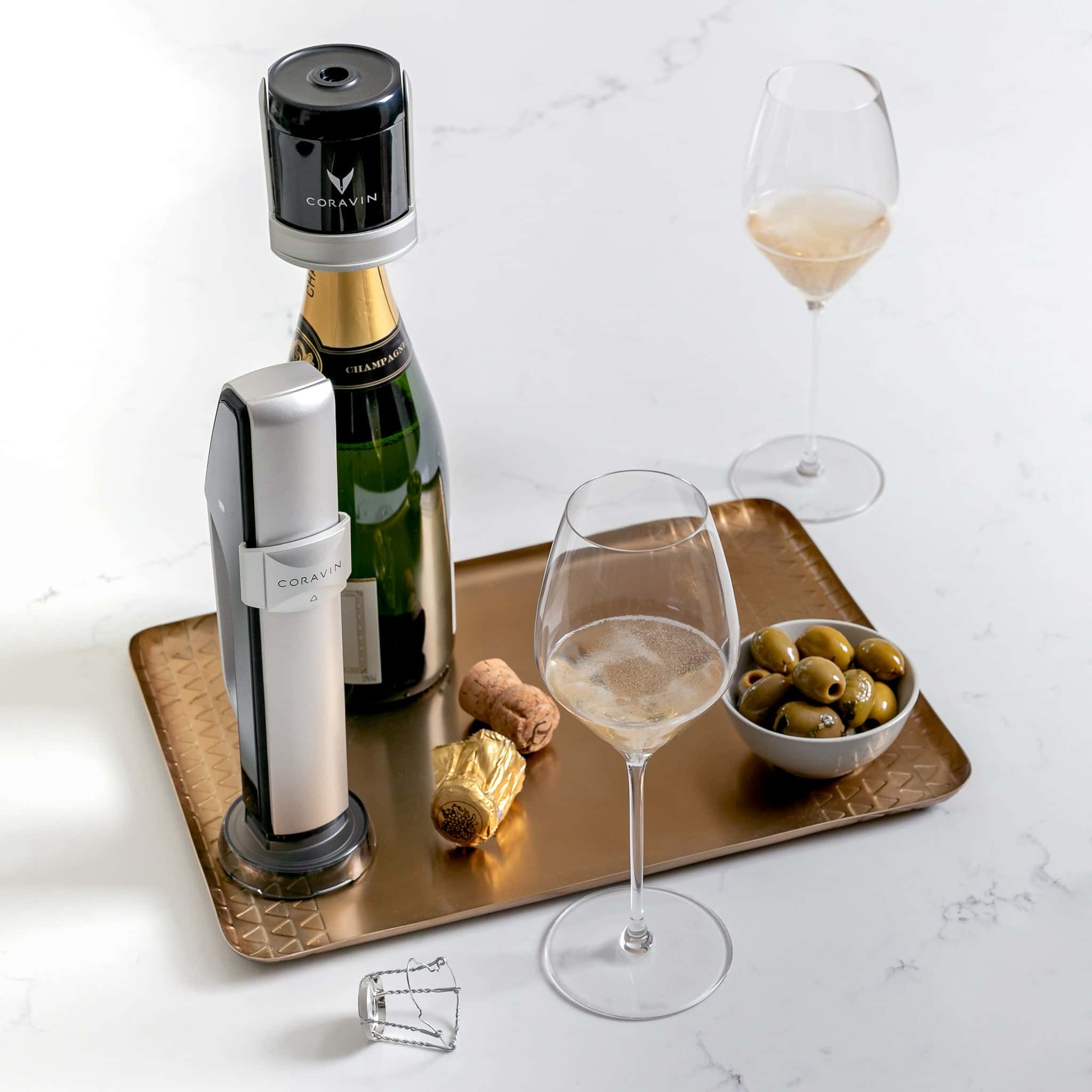 Coravin Sparkling Wine Preservation System with a champagne bottle, two flutes of sparkling wine, green olives, and cork on a gold tray over a marble surface.