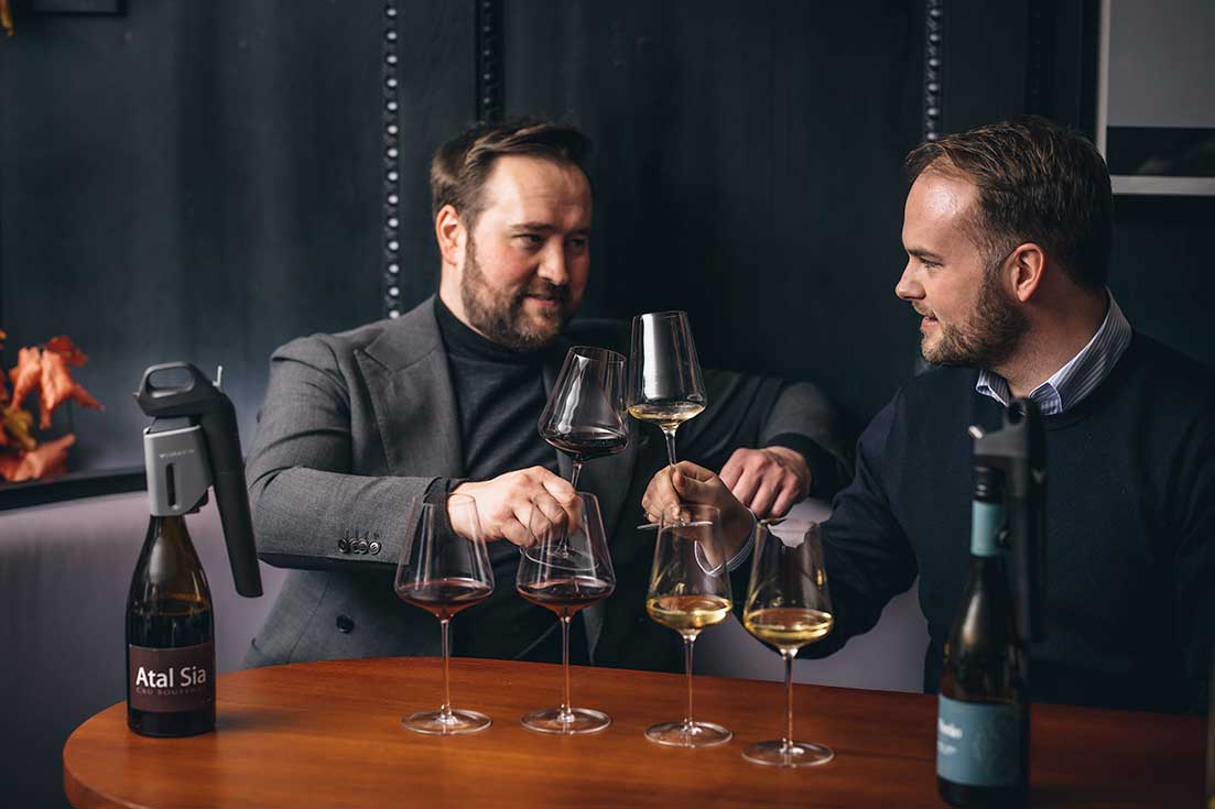 Two men clinking wine glasses at a tasting table with red and white wines, each bottle equipped with a Coravin wine preservation system