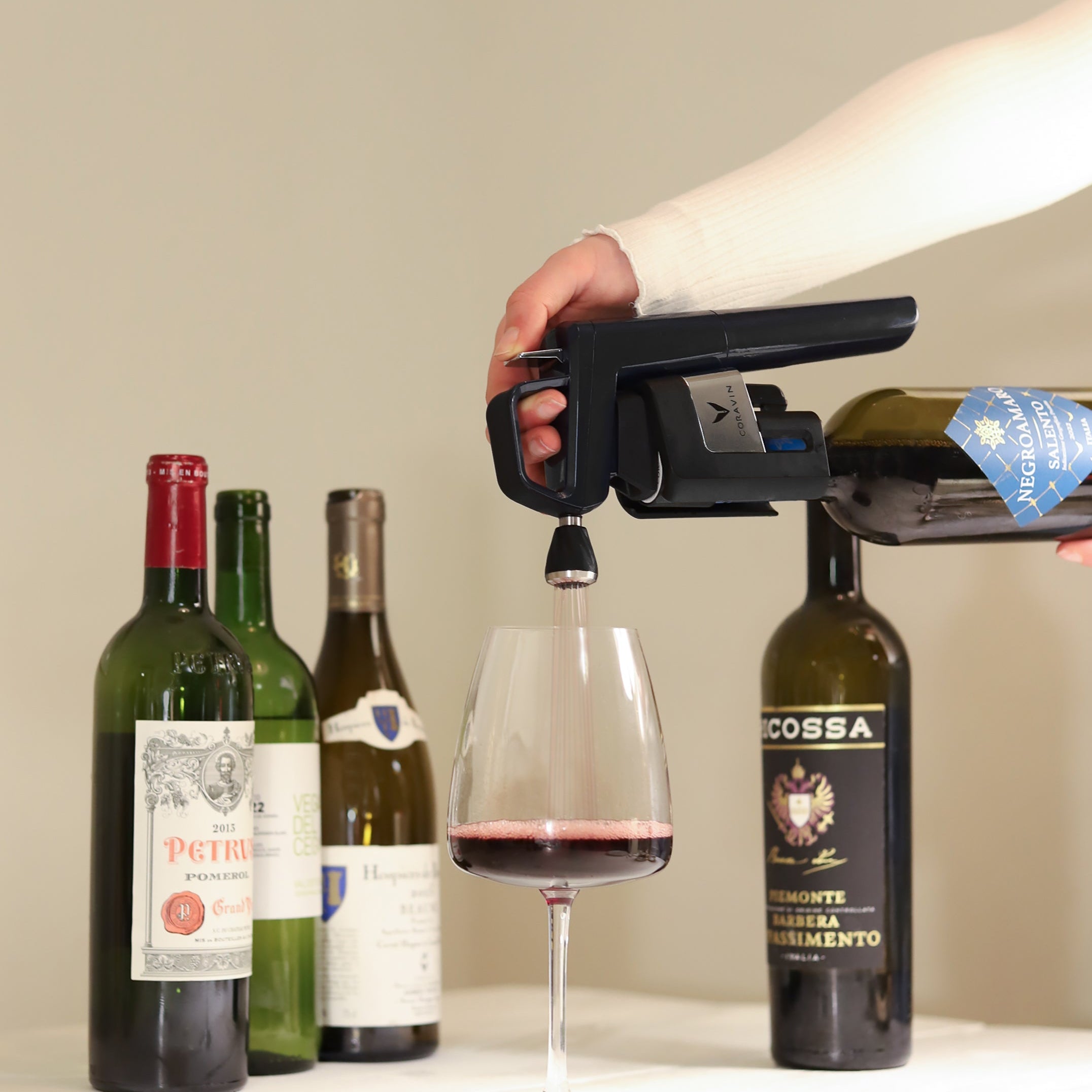 Person using a Coravin Timeless wine preservation system to pour red wine from a bottle into a glass, with several premium wine bottles displayed in the background.