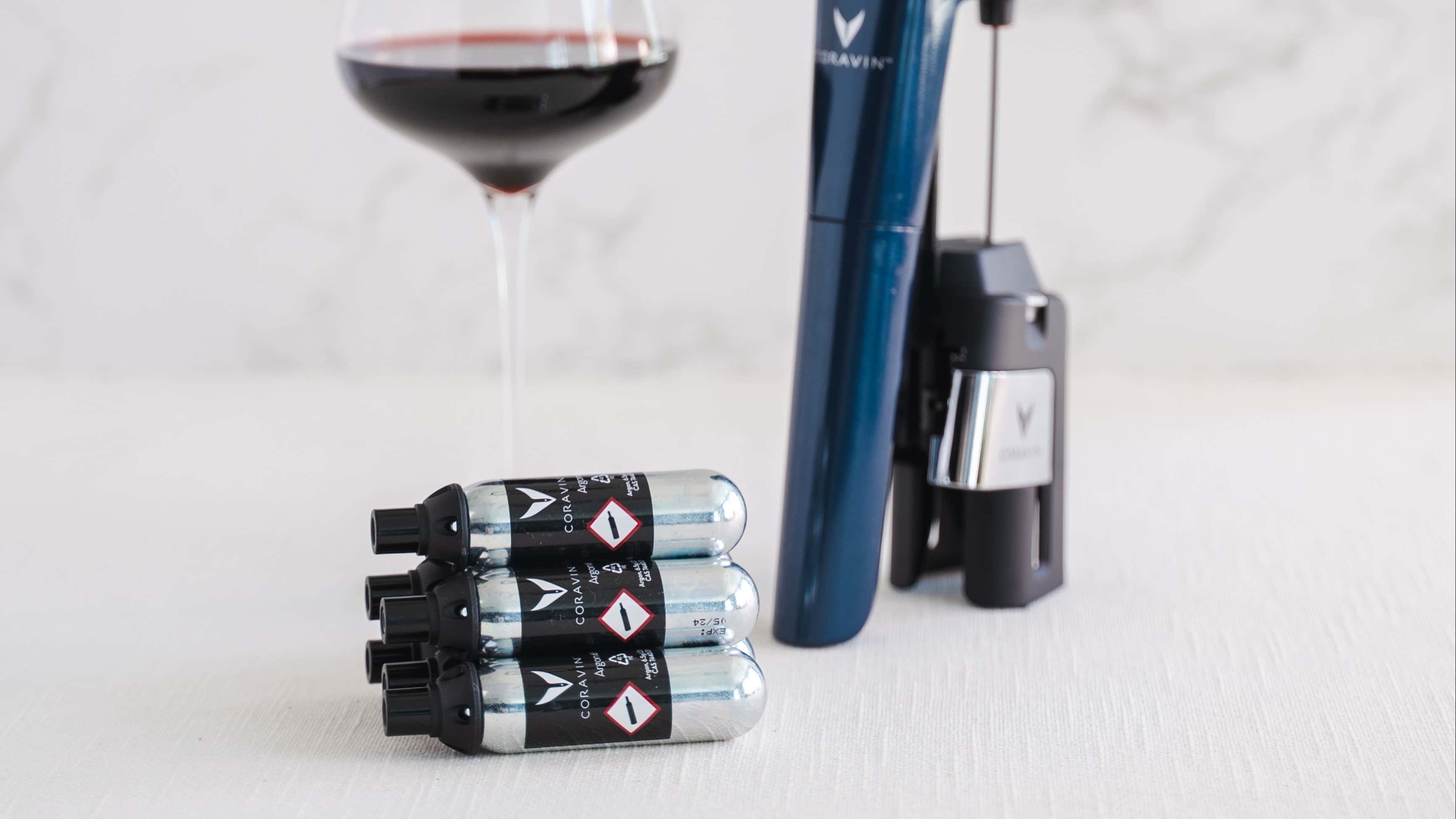 Three Coravin Pure argon capsules stacked on a light-colored surface in front of a Coravin wine-preserving device and a partially visible glass of red wine, all set against a soft, marble-patterned background