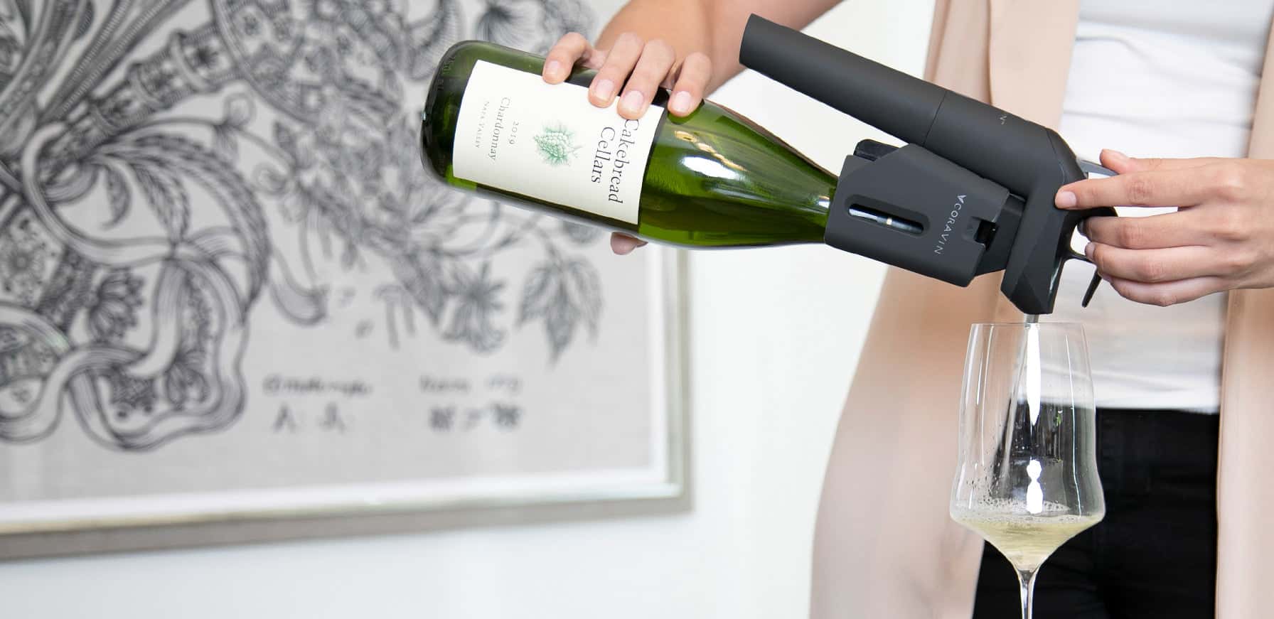Person using Coravin wine preservation system to pour Chardonnay from a Cakebread Cellars bottle into a glass, with modern art in the background.