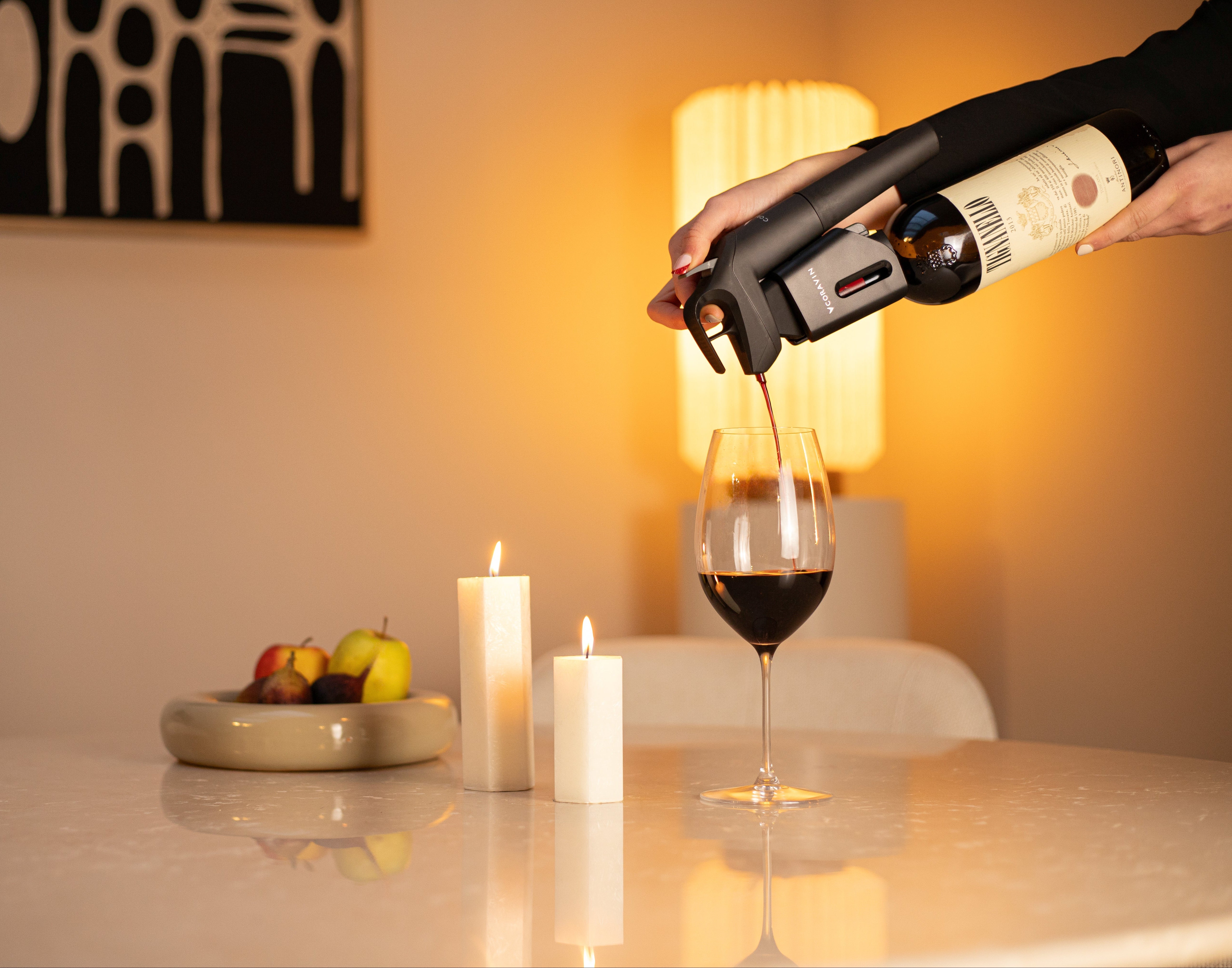 Hand pouring red wine using a Coravin wine preservation system into a glass on a modern candlelit dining table, with warm ambient lighting and a bowl of fruit in the background.