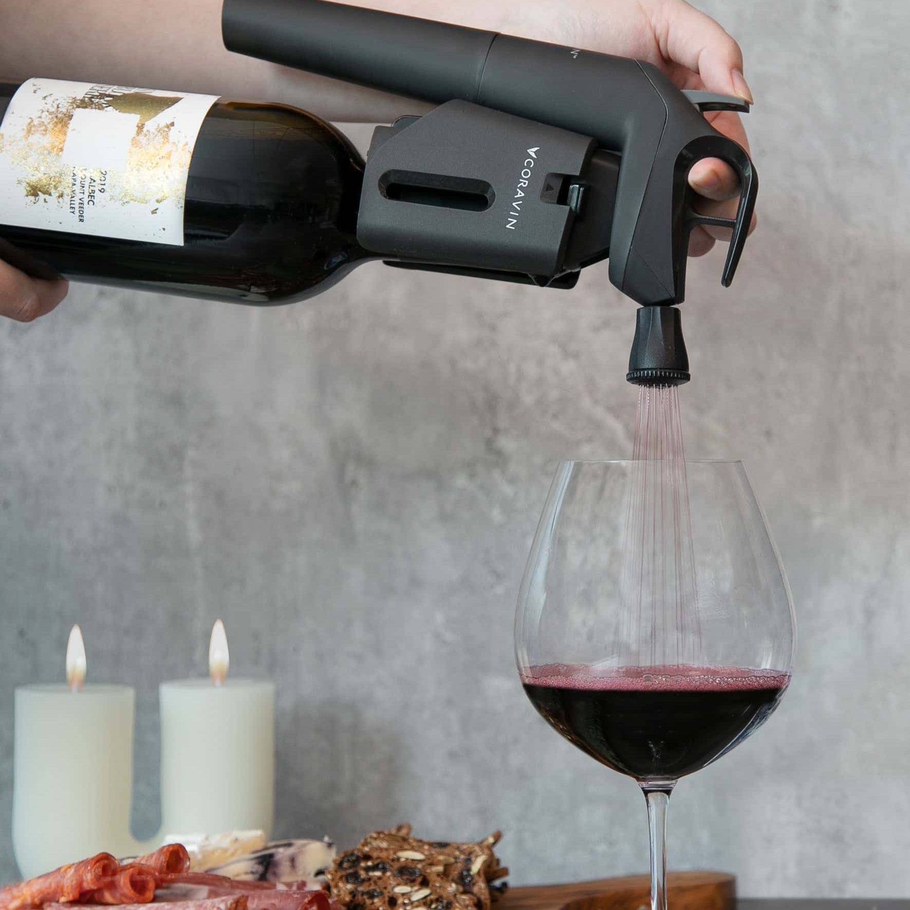 Pouring red wine using Coravin Timeless Three+ wine preservation system, with a charcuterie board and candles in the background.