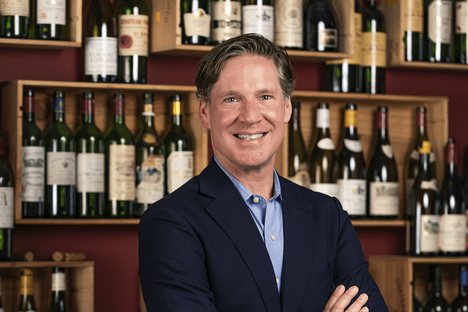 Greg Lambrecht, founder of Coravin, smiling in front of a wall of wine bottles, showcasing his dedication to wine preservation and innovation.