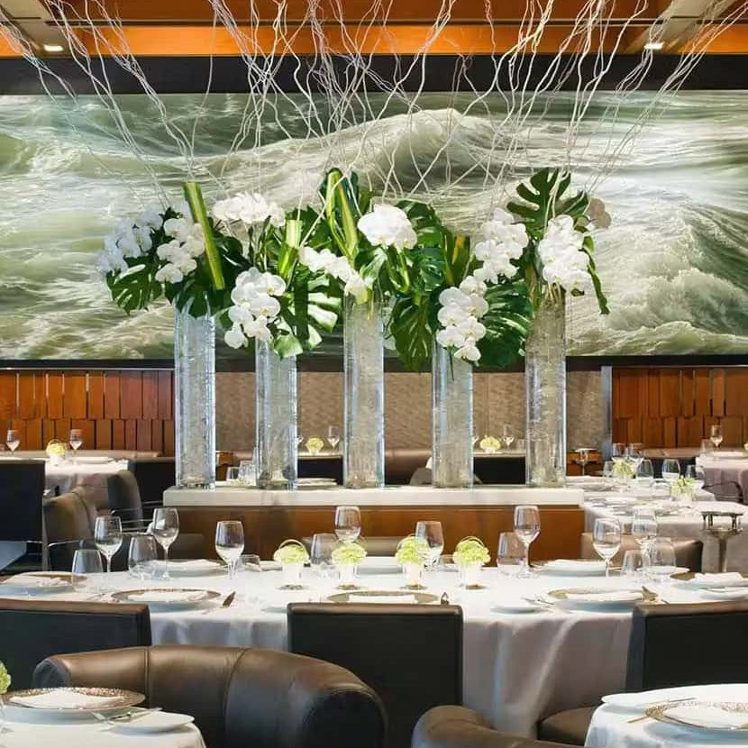 Elegant fine dining restaurant with round white tablecloth tables, modern leather chairs, and tall glass vases filled with white orchids and tropical greenery.