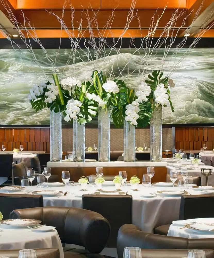 Elegant fine dining restaurant with round white tablecloth tables, modern leather chairs, and tall glass vases filled with white orchids and tropical greenery.