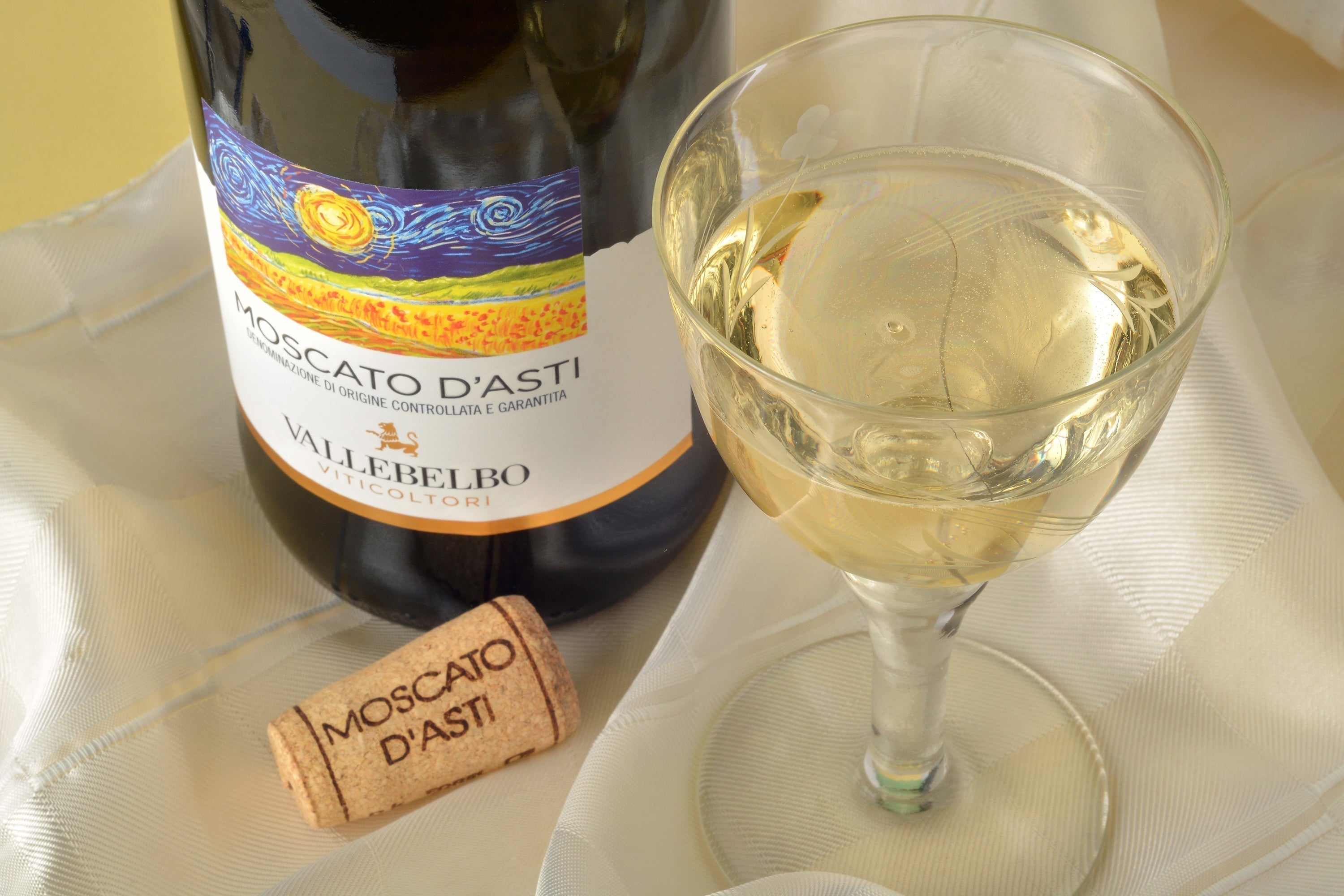 What Is Moscato: A close-up of a glass of golden Moscato d’Asti wine beside its bottle and cork, set on a soft cream fabric background.