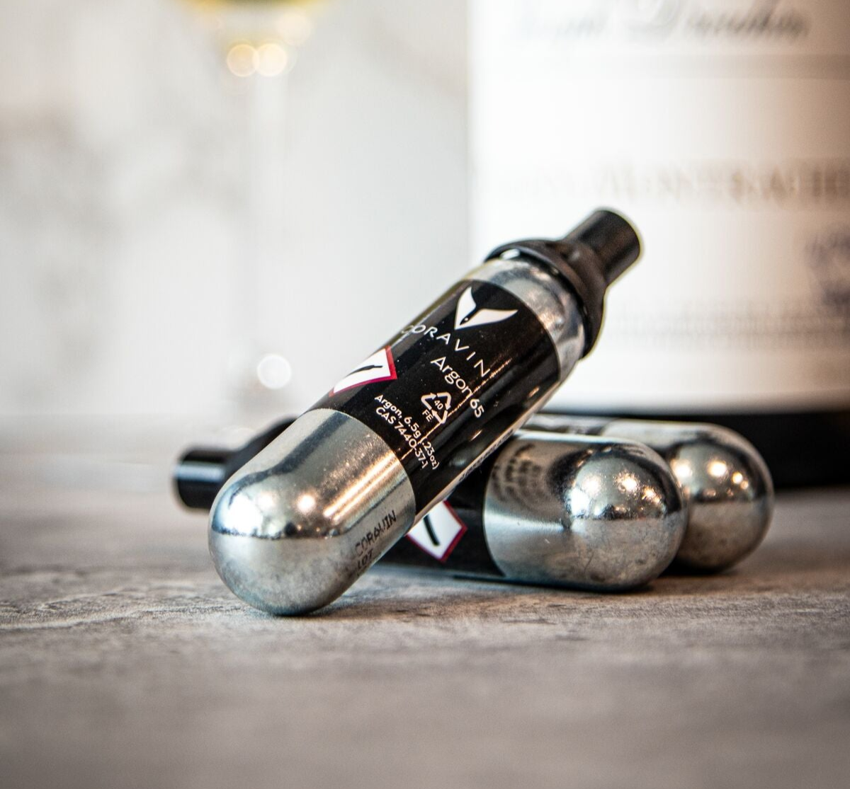 Three silver gas with black labels are arranged on a textured surface, with a blurred wine bottle in the background.