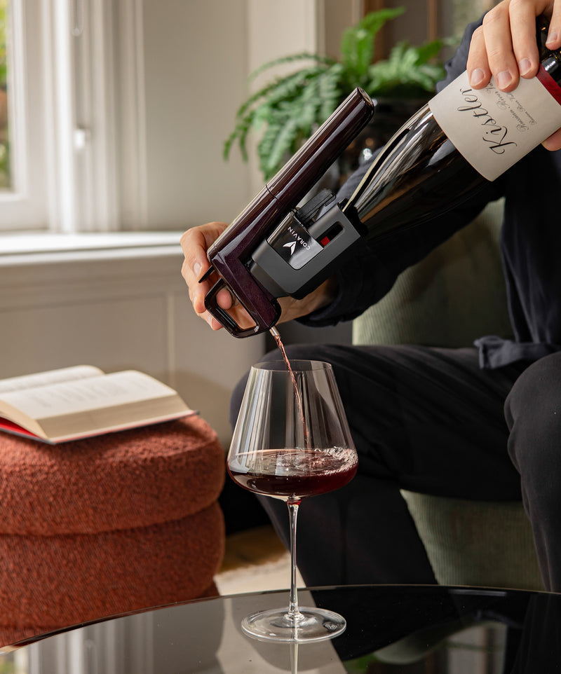 Person pouring red wine from a bottle with coravin timeless six into a glass in a cozy indoor setting.