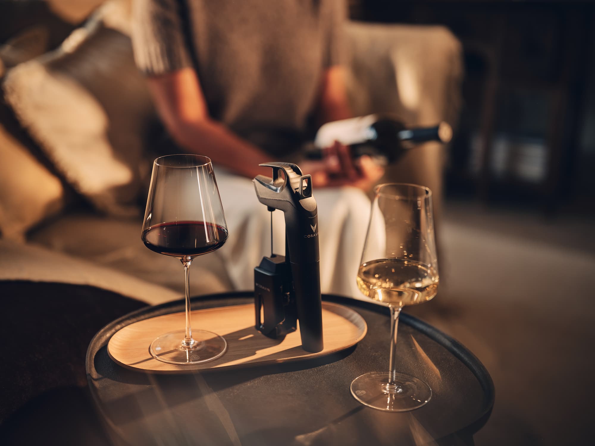 Coravin wine system placed between two wine glasses—one with red wine, one with white—on a small round table with a wooden tray. Refined home atmosphere