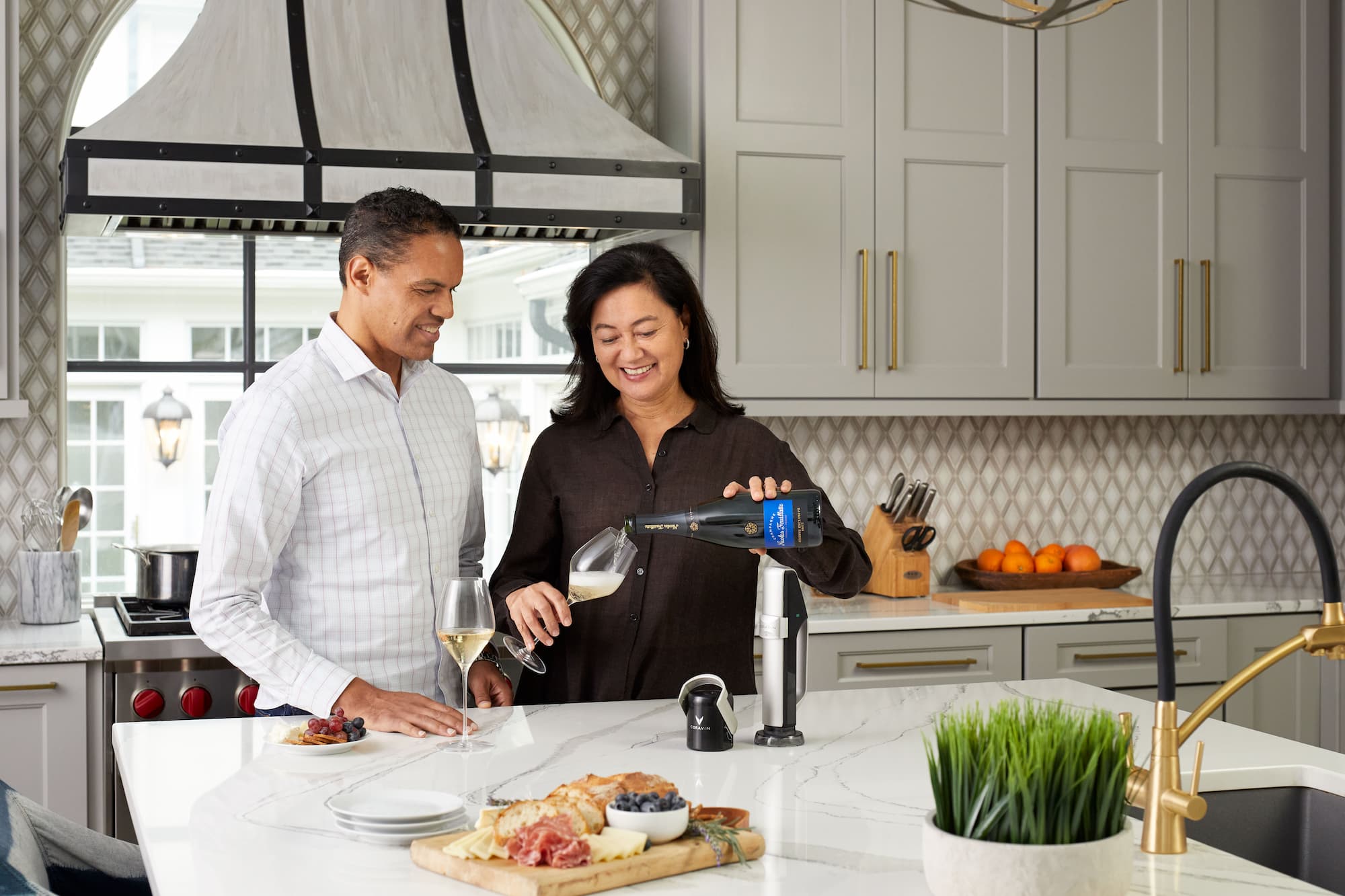 Two people enjoying wine and charcuterie in a modern kitchen; one pours sparkling wine using a Coravin system