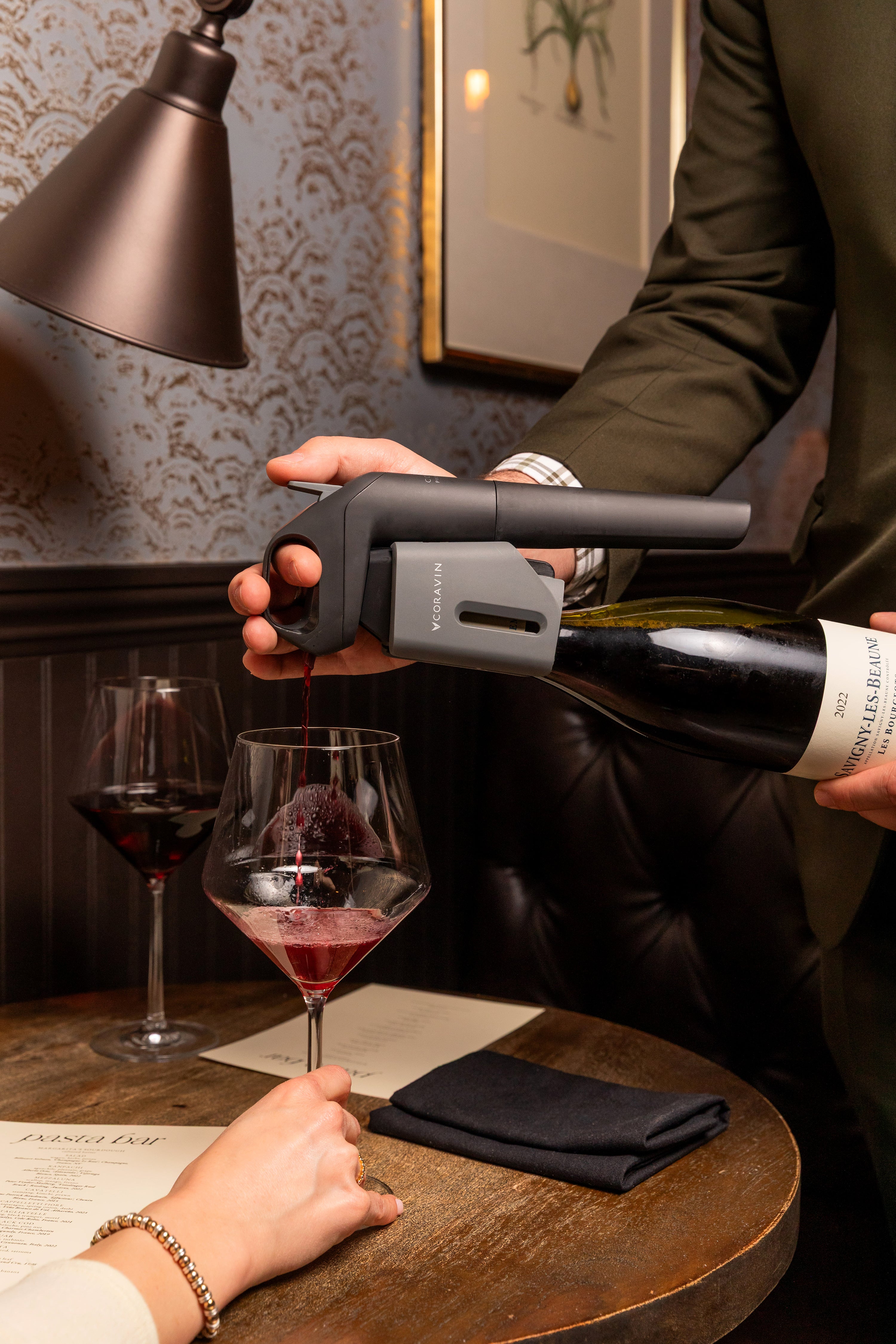 A person pours red wine using a Coravin Timeless Six+™ wine by-the-glass system into a glass held by a guest at Pasta | Bar. A tasting menu lies on the table.