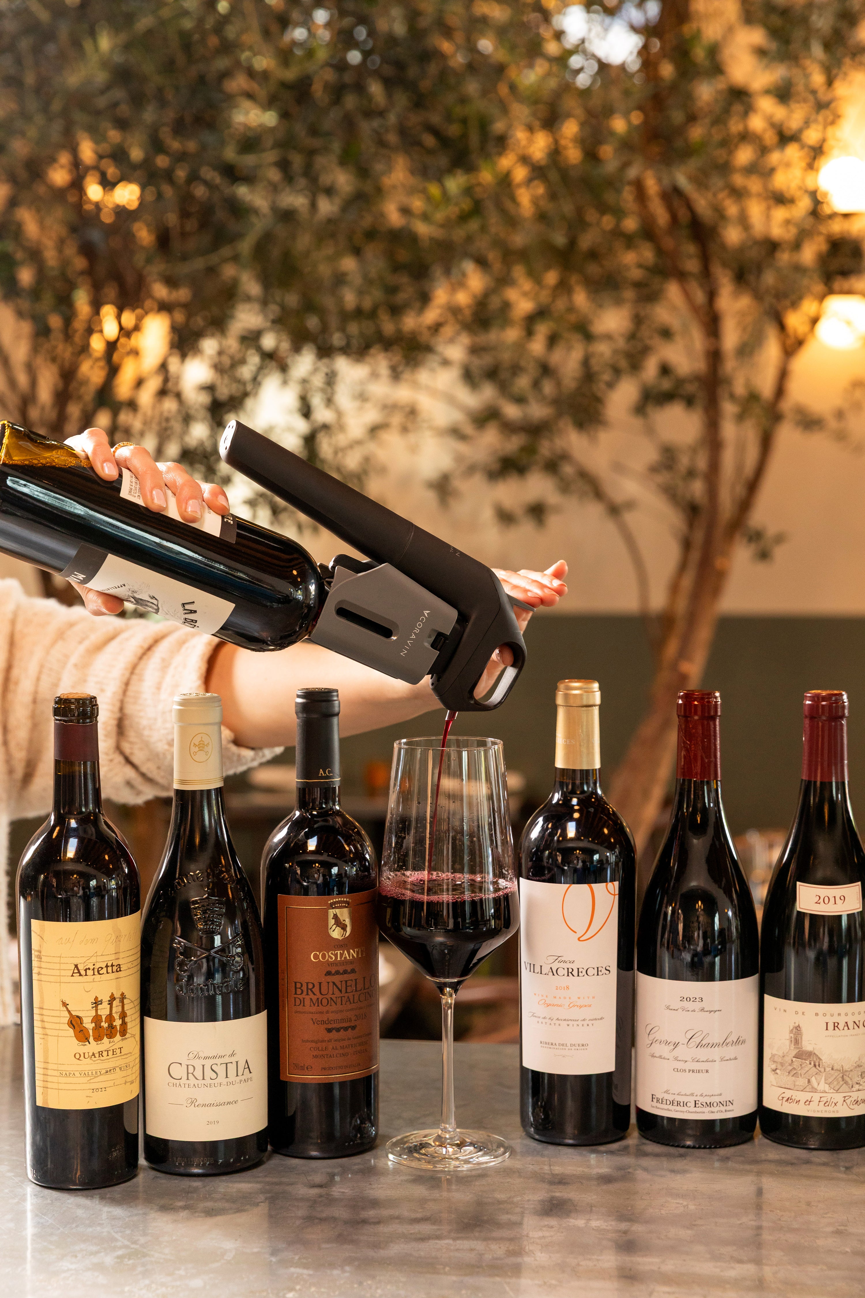 Pouring red wine using a Coravin Timeless Six+™ wine by-the-glass system at a wine bar, surrounded by premium red wine bottles including Brunello, Châteauneuf-du-Pape, and Napa Valley wines.